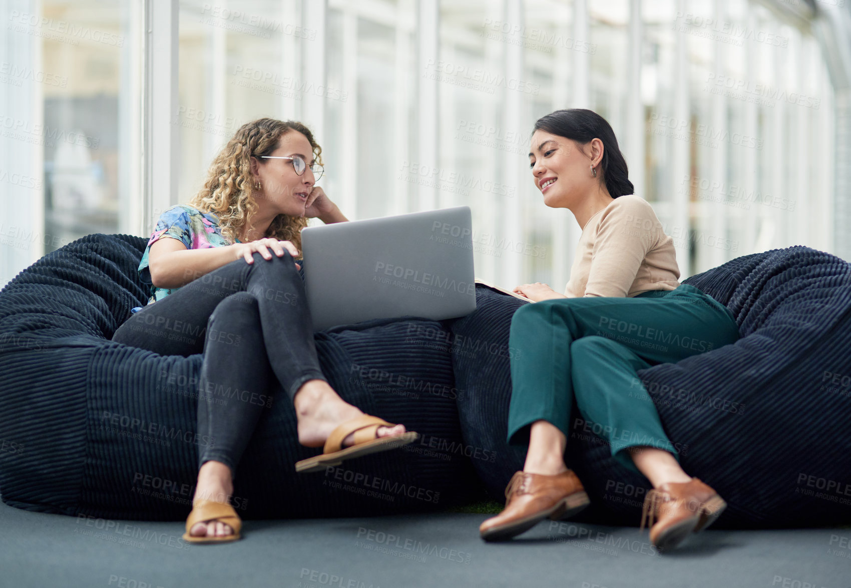 Buy stock photo Shot of two businesswomen using a laptop together while sitting on beanbags in an office