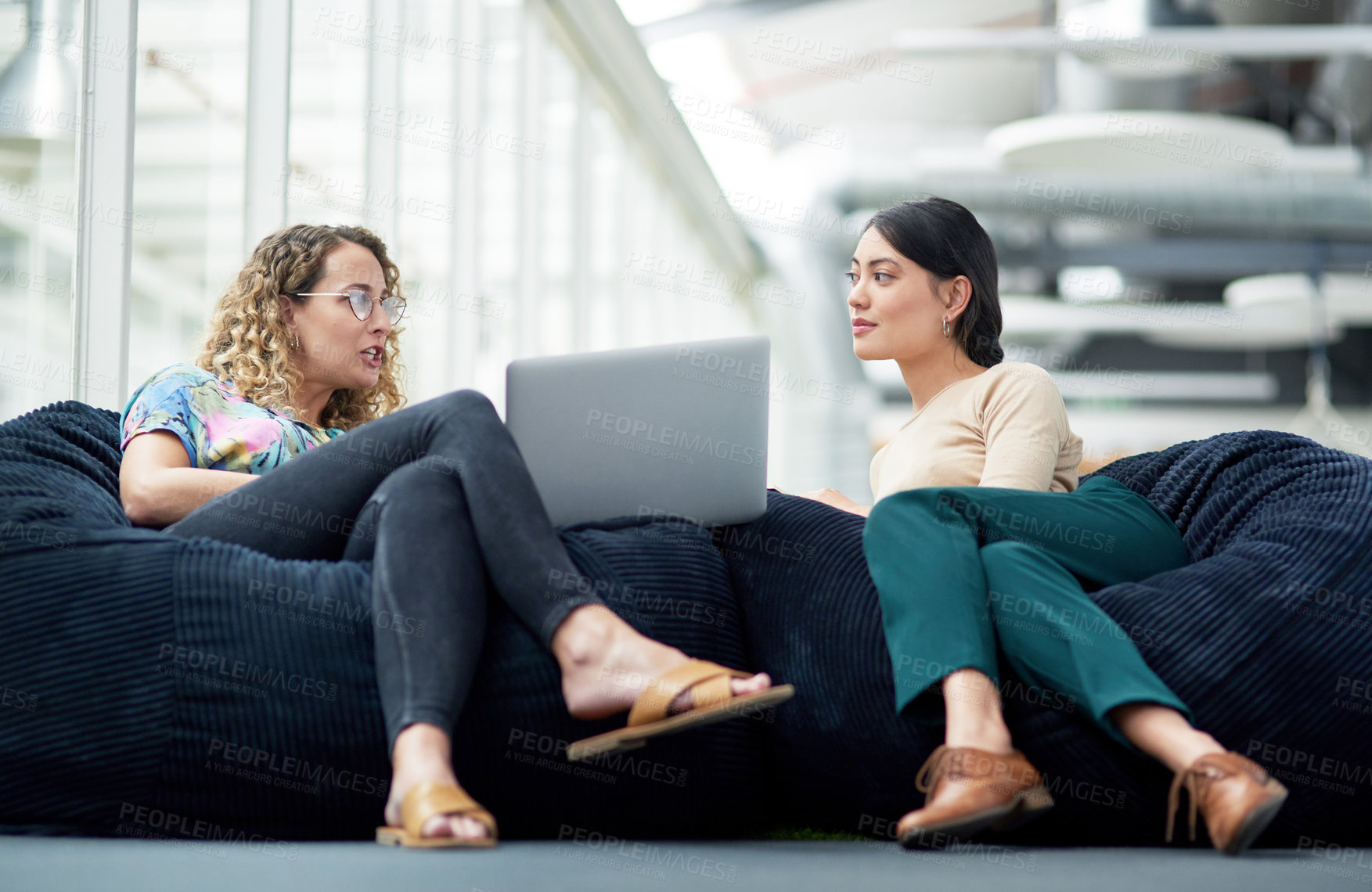 Buy stock photo Shot of two businesswomen using a laptop together while sitting on beanbags in an office