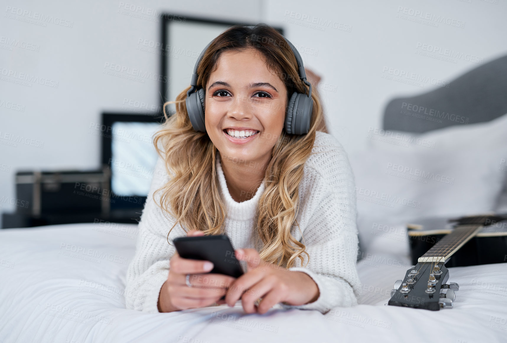 Buy stock photo Shot of a young woman wearing headphones and holding her cellphone while lying on her bed