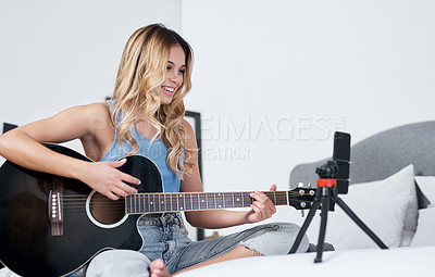 Buy stock photo Shot of a young woman using her cellphone to record herself while playing the guitar at home