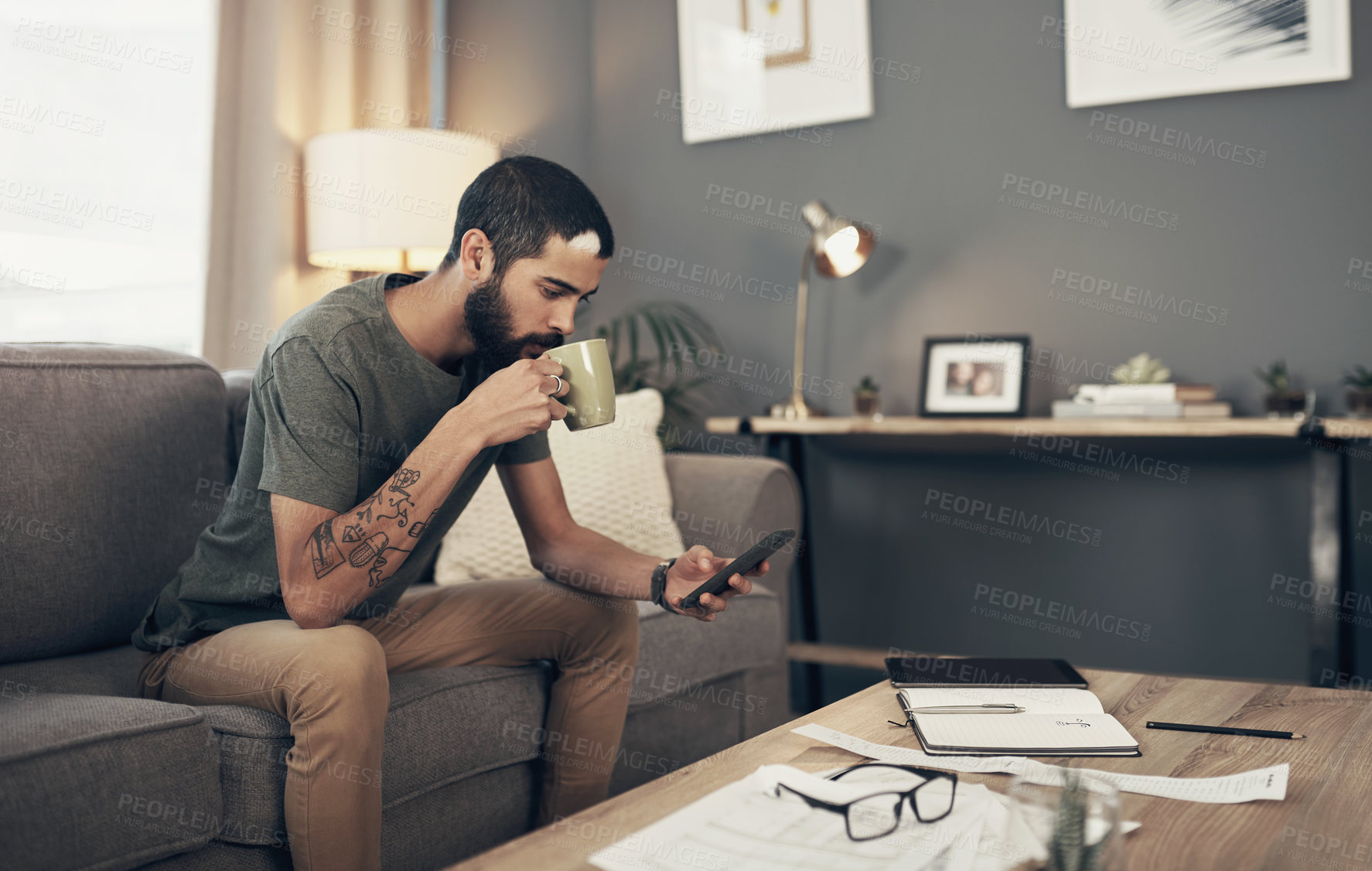 Buy stock photo Shot of a young man having coffee and using a smartphone while going through paperwork at home