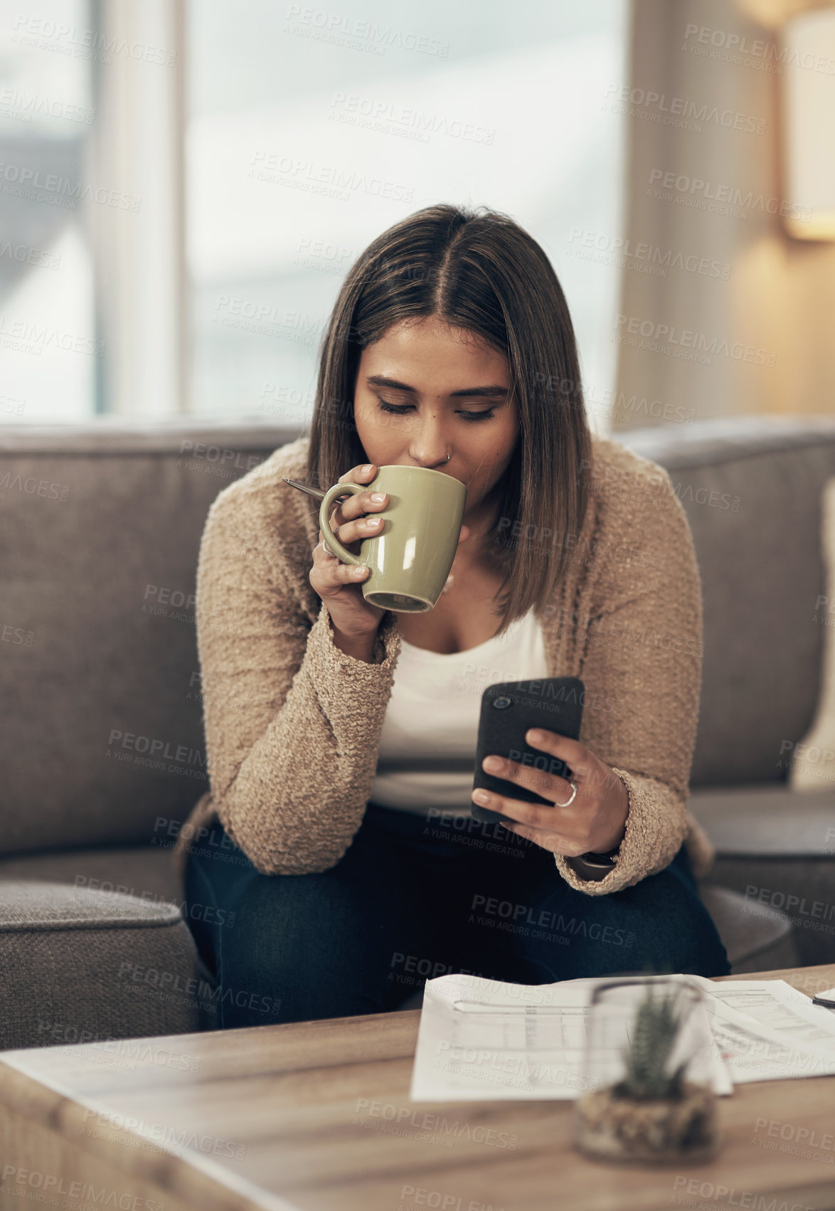 Buy stock photo Shot of a young woman having coffee and using a smartphone while going through paperwork at home
