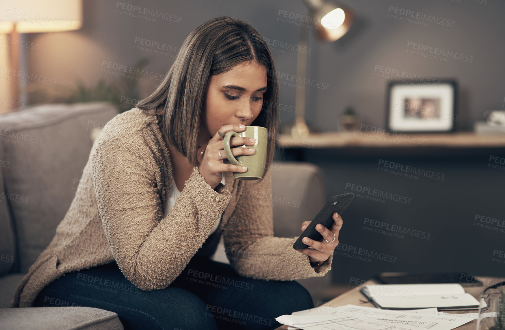 Buy stock photo Shot of a young woman having coffee and using a smartphone while going through paperwork at home
