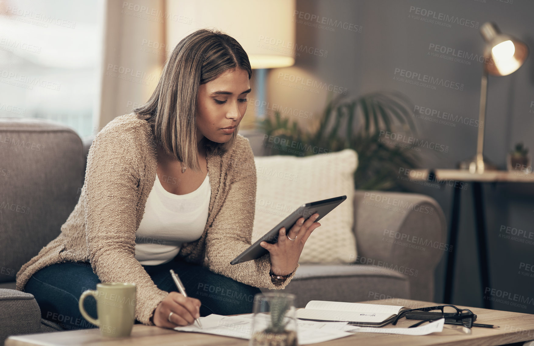 Buy stock photo Shot of a young woman using a digital tablet while going through paperwork at home
