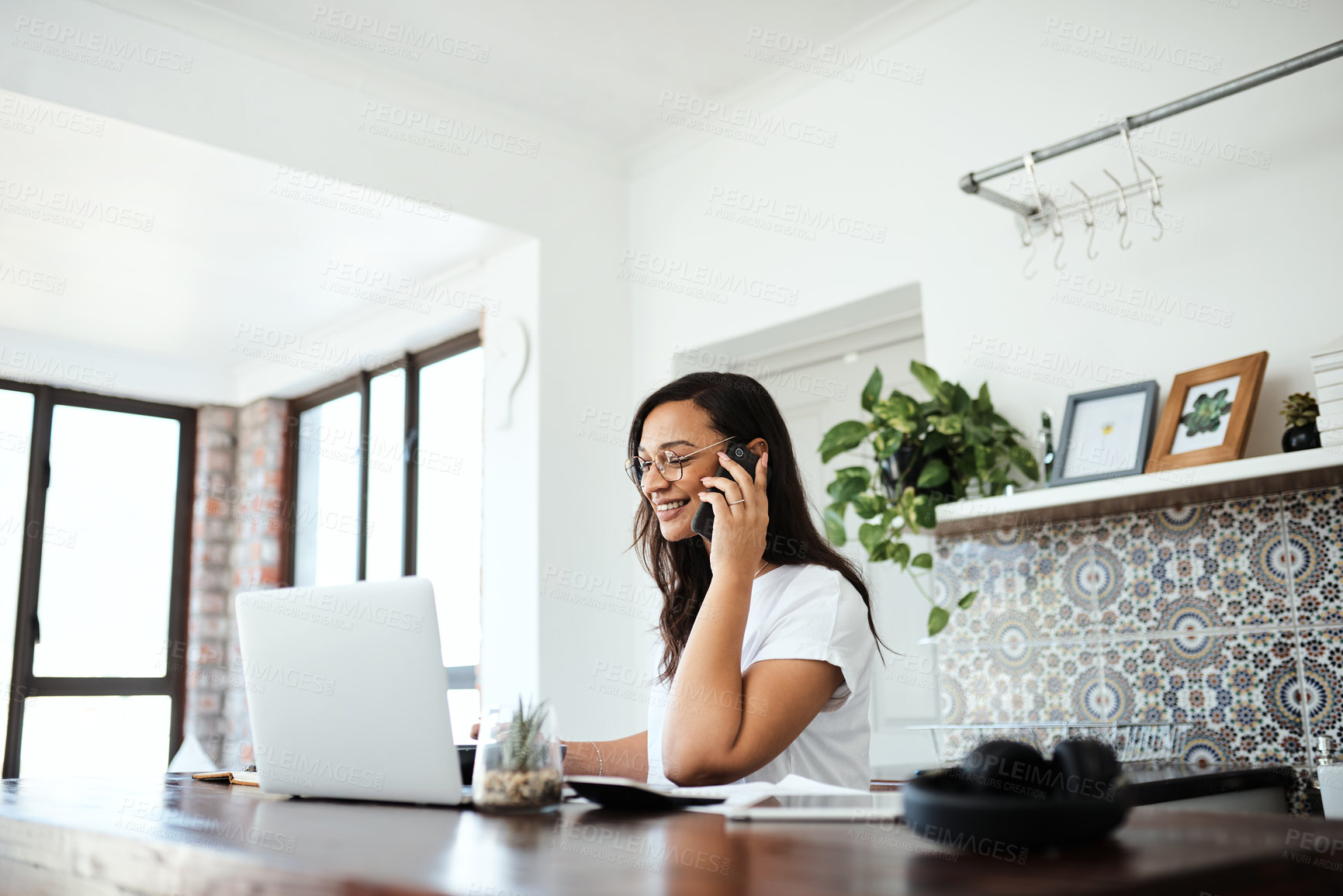 Buy stock photo Shot of a young woman talking on a cellphone while working on a laptop at home