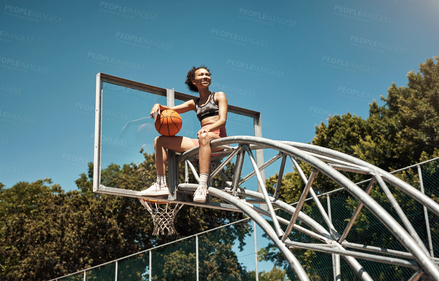 Buy stock photo Shot of a sporty young woman sitting on a basketball hoop on a sports court