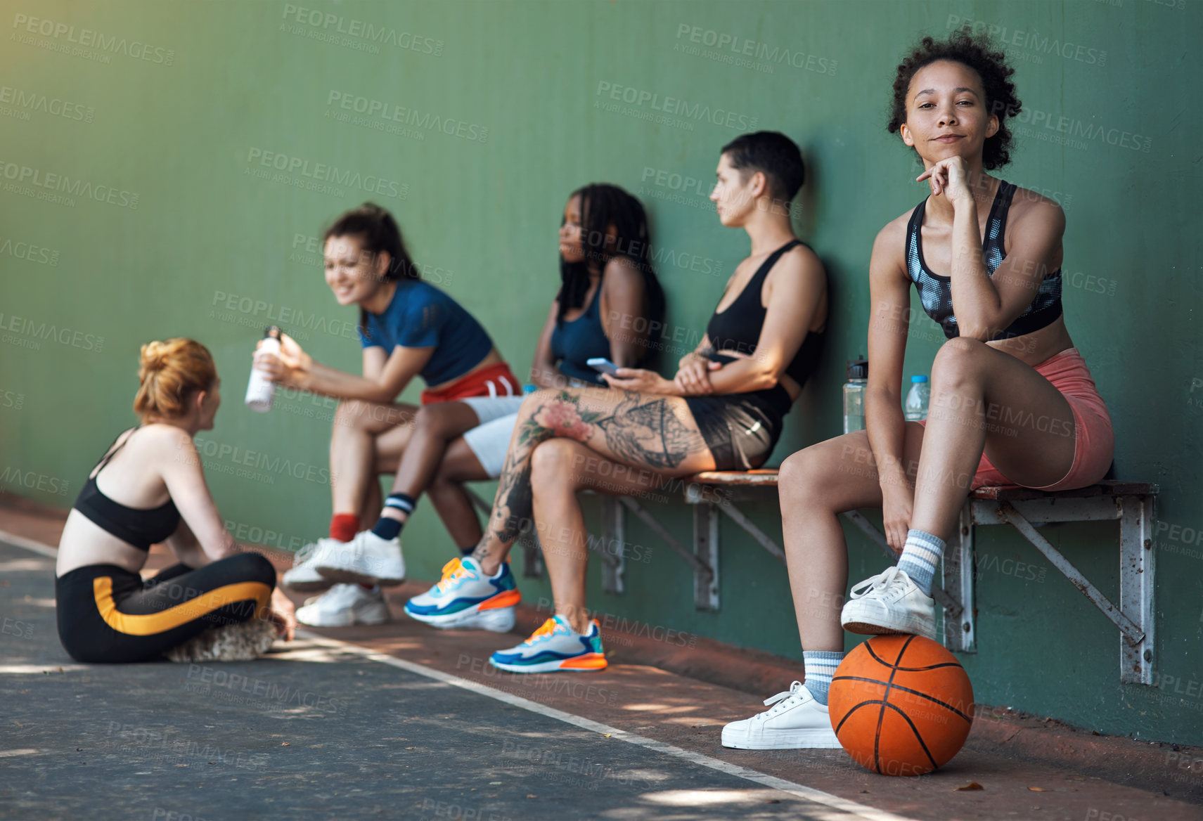 Buy stock photo Full length portrait of an attractive young female athlete sitting on a bench at the basketball court with her teammates in the background