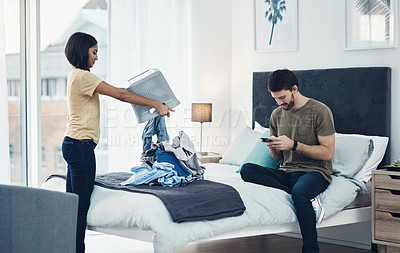 Buy stock photo Shot of a young woman doing laundry while her husband uses his smartphone at home