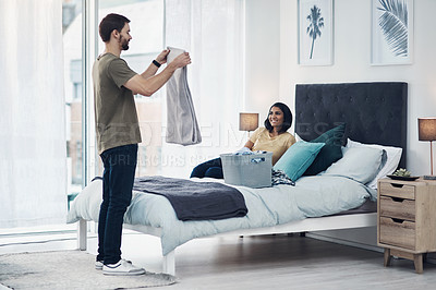 Buy stock photo Shot of a young man folding laundry while his wife relaxes on the bed at home