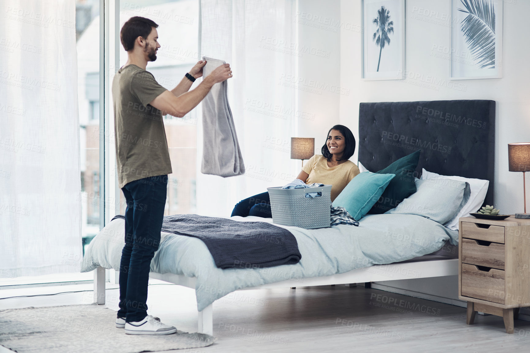 Buy stock photo Shot of a young man folding laundry while his wife relaxes on the bed at home