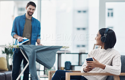 Buy stock photo Shot of a young woman using a smartphone and headphones while her husband irons clothing in the background