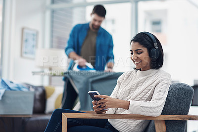 Buy stock photo Shot of a young woman using a smartphone and headphones while her husband irons clothing in the background