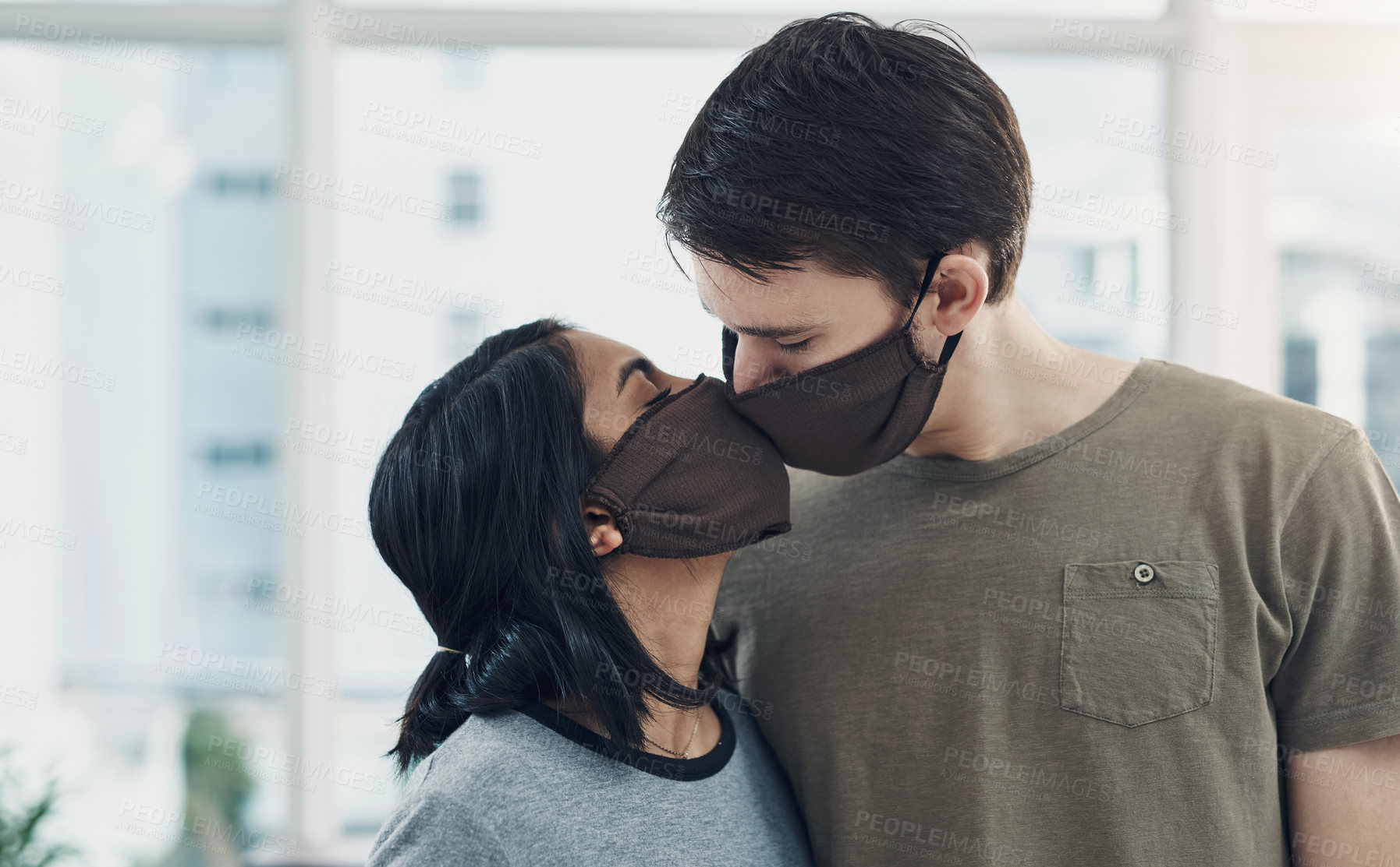 Buy stock photo Shot of a young couple kissing each other while wearing masks at home