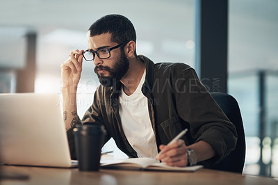 Buy stock photo Shot of a young businessman writing in a notebook and using a laptop during a late night in a modern office