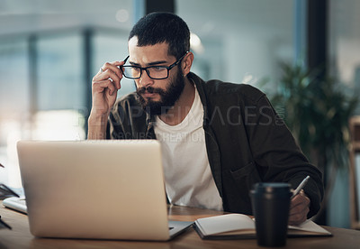 Buy stock photo Shot of a young businessman writing in a notebook and using a laptop during a late night in a modern office