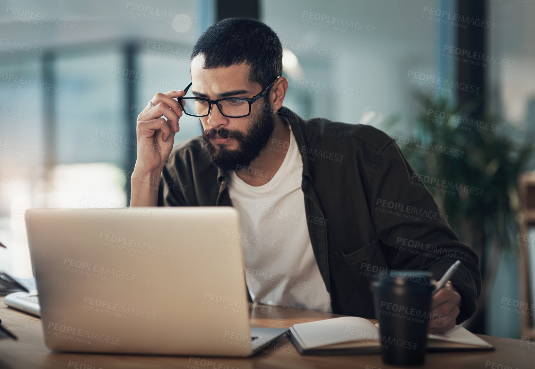 Buy stock photo Shot of a young businessman writing in a notebook and using a laptop during a late night in a modern office