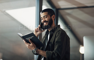 Buy stock photo Shot of a young businessman using a smartphone and reading a notebook during a late night in a modern office