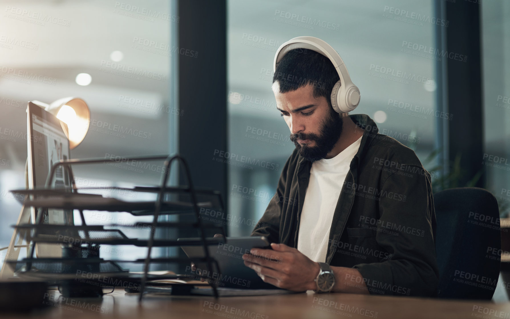 Buy stock photo Shot of a young businessman using a digital tablet with headphones during a late night at work