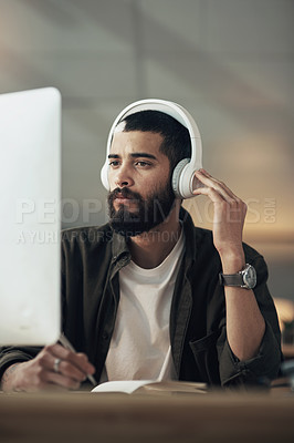 Buy stock photo Shot of a young businessman writing in a notebook and using a computer with headphones during a late night at work