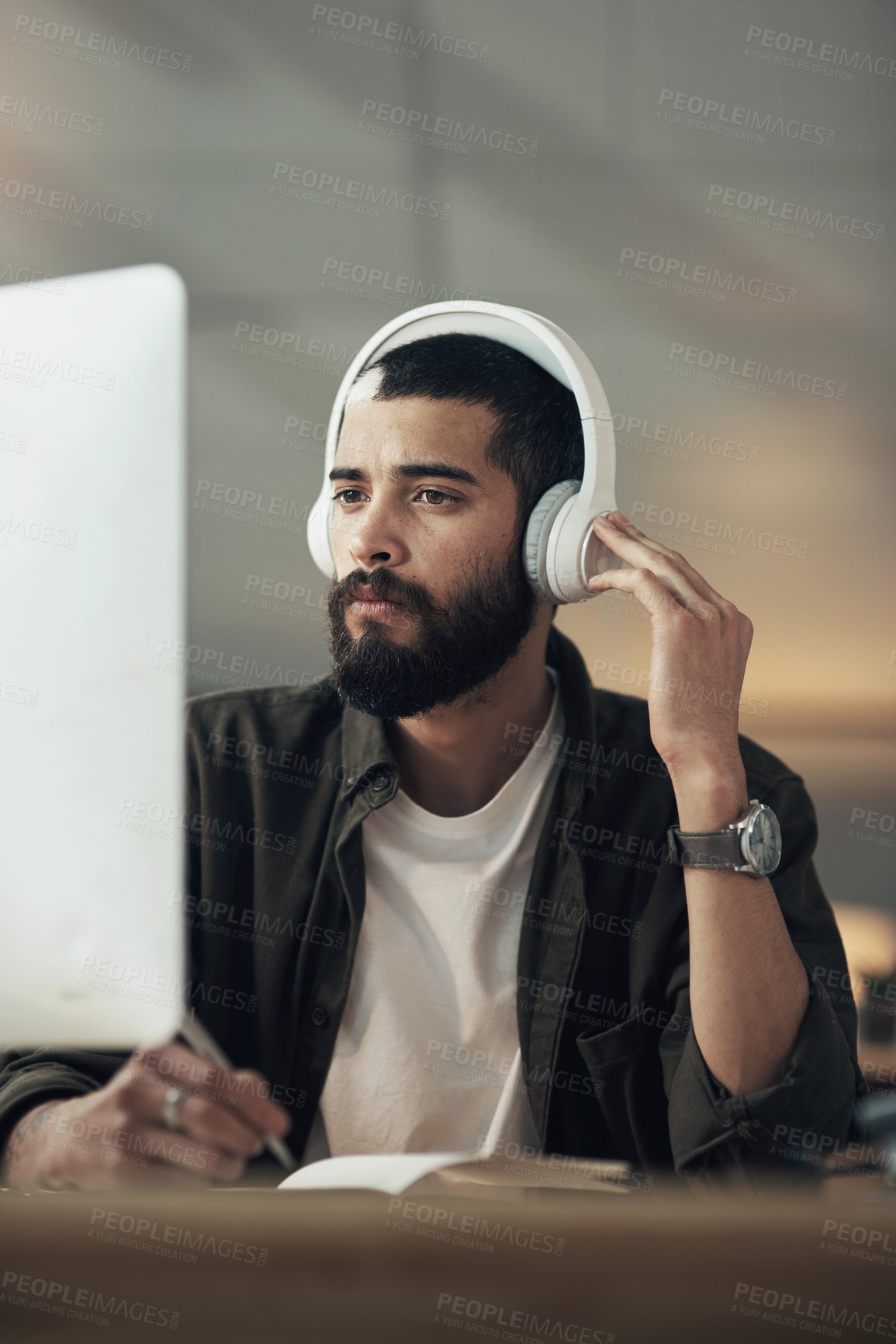 Buy stock photo Shot of a young businessman writing in a notebook and using a computer with headphones during a late night at work