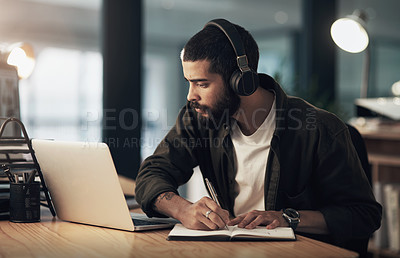 Buy stock photo Shot of a young businessman writing in a notebook and using a laptop with headphones during a late night at work