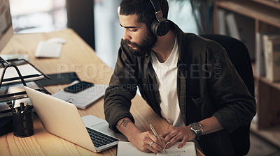 Buy stock photo Shot of a young businessman writing in a notebook and using a laptop with headphones during a late night at work
