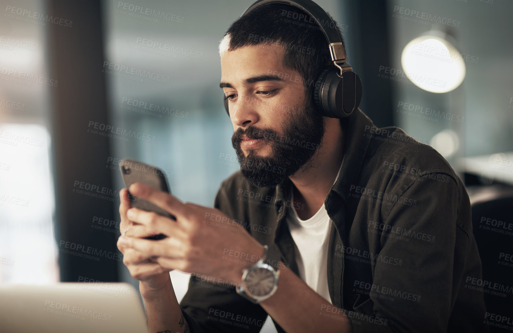 Buy stock photo Shot of a young businessman using a smartphone with headphones during a late night at work