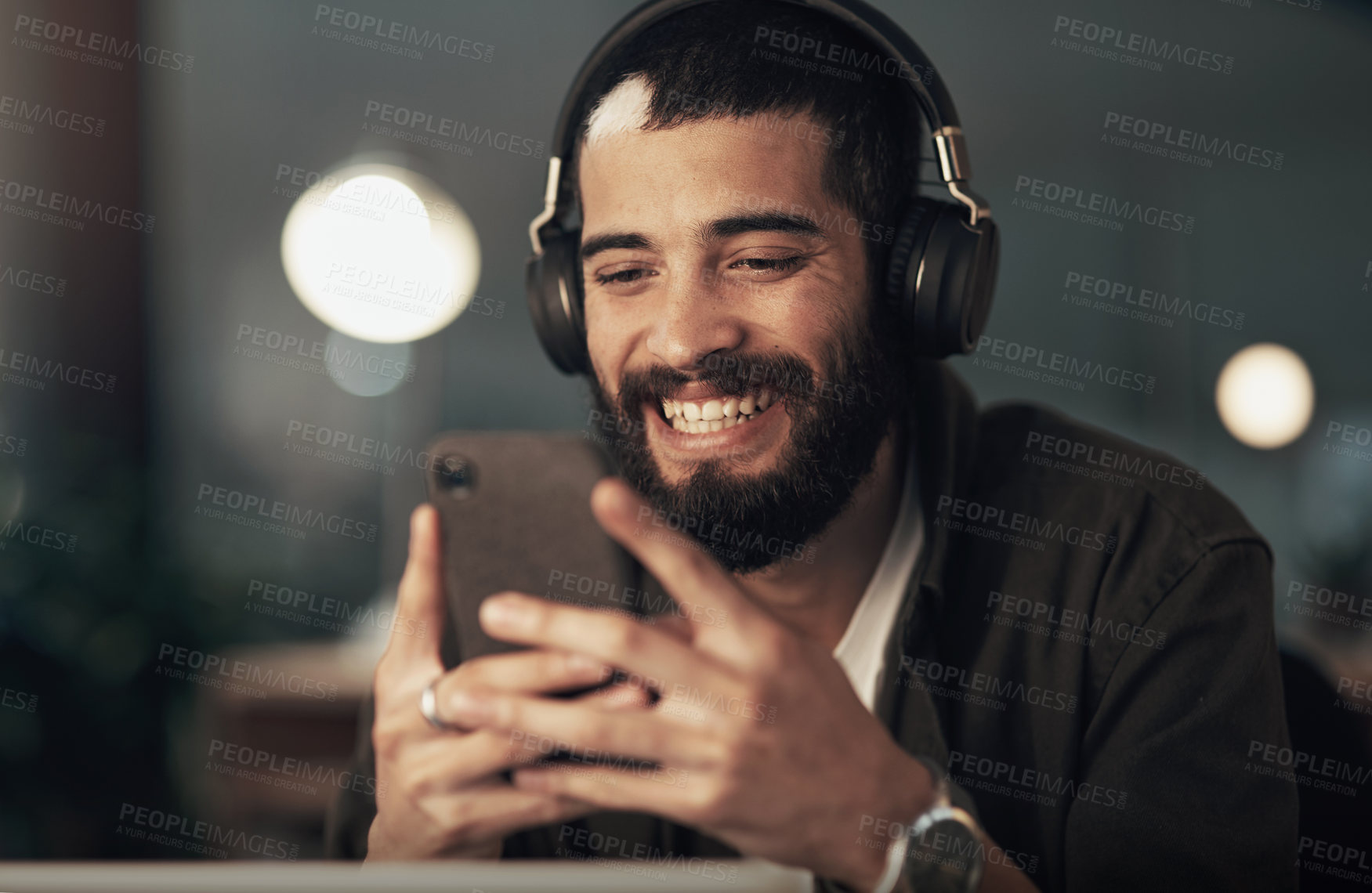 Buy stock photo Shot of a young businessman using a smartphone with headphones during a late night at work