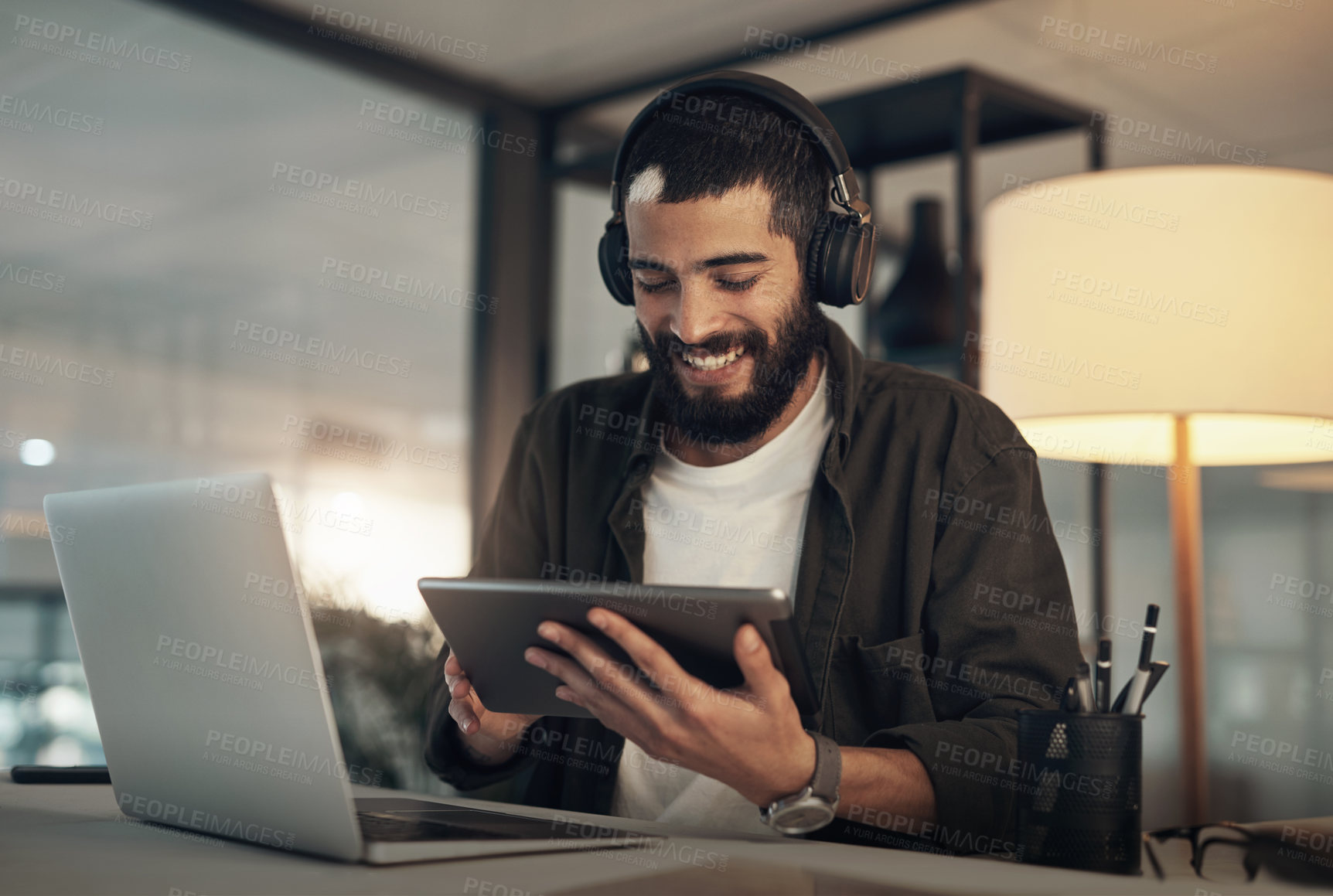 Buy stock photo Shot of a young businessman using a digital tablet, laptop and headphones during a late night at work