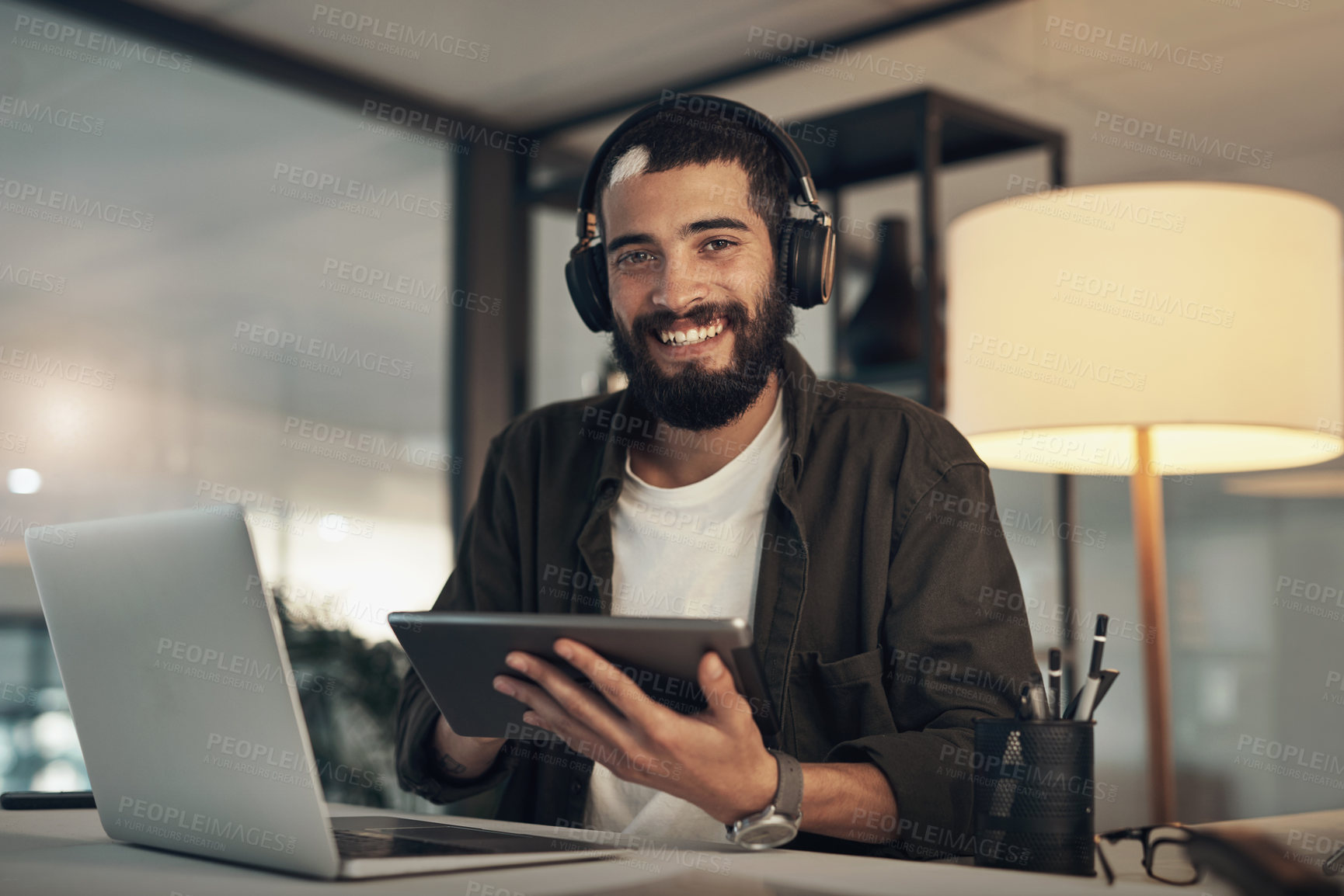 Buy stock photo Shot of a young businessman using a digital tablet, laptop and headphones during a late night at work