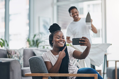 Buy stock photo Shot of a young woman taking selfies while her husband irons clothing in the background