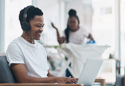 Buy stock photo Shot of a young man using a laptop while his wife irons clothing in the background