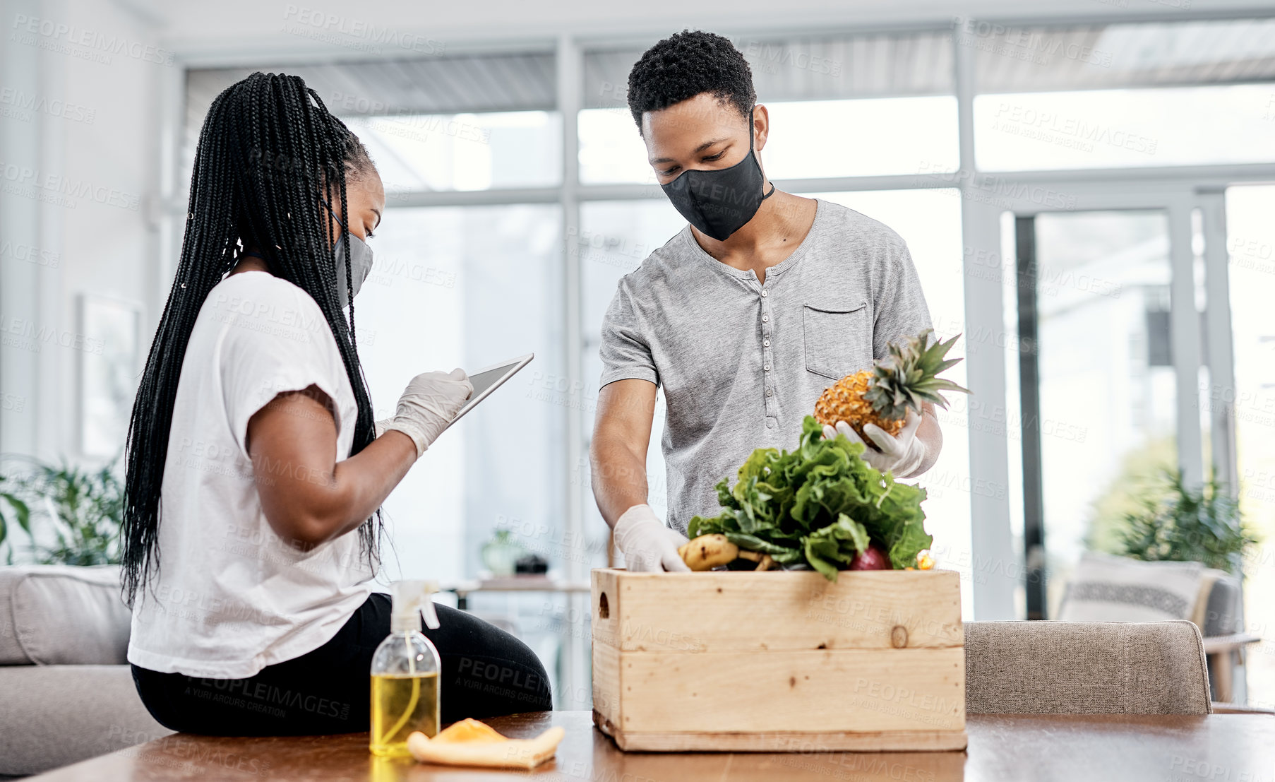 Buy stock photo Shot of a masked young couple using a digital tablet while disinfecting their groceries at home