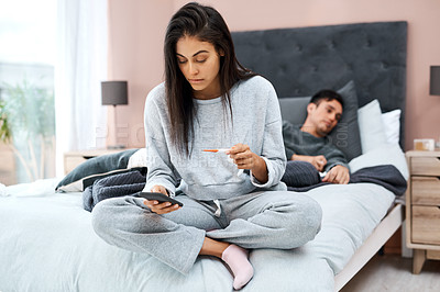 Buy stock photo Shot of a young woman using a smartphone and thermometer while recovering from an illness with her husband at home