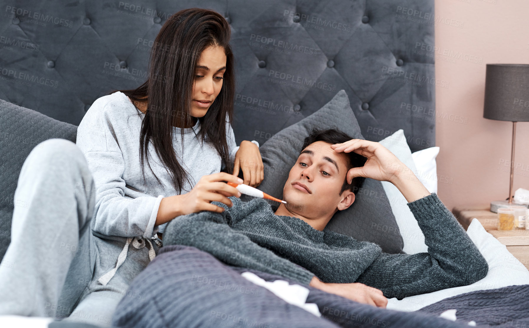 Buy stock photo Shot of a young woman taking her husband’s temperature while recovering from an illness in bed at home
