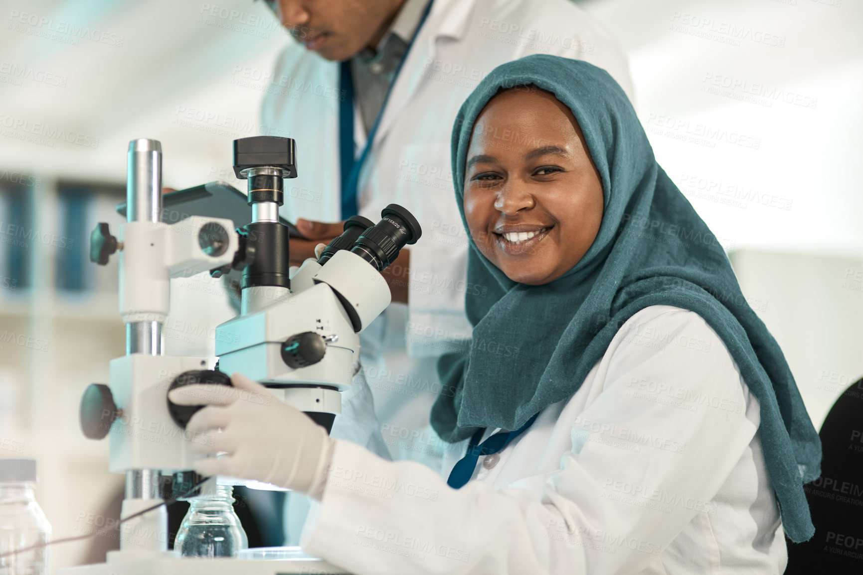Buy stock photo Portrait of a young scientist using a microscope while working alongside a colleague in a lab