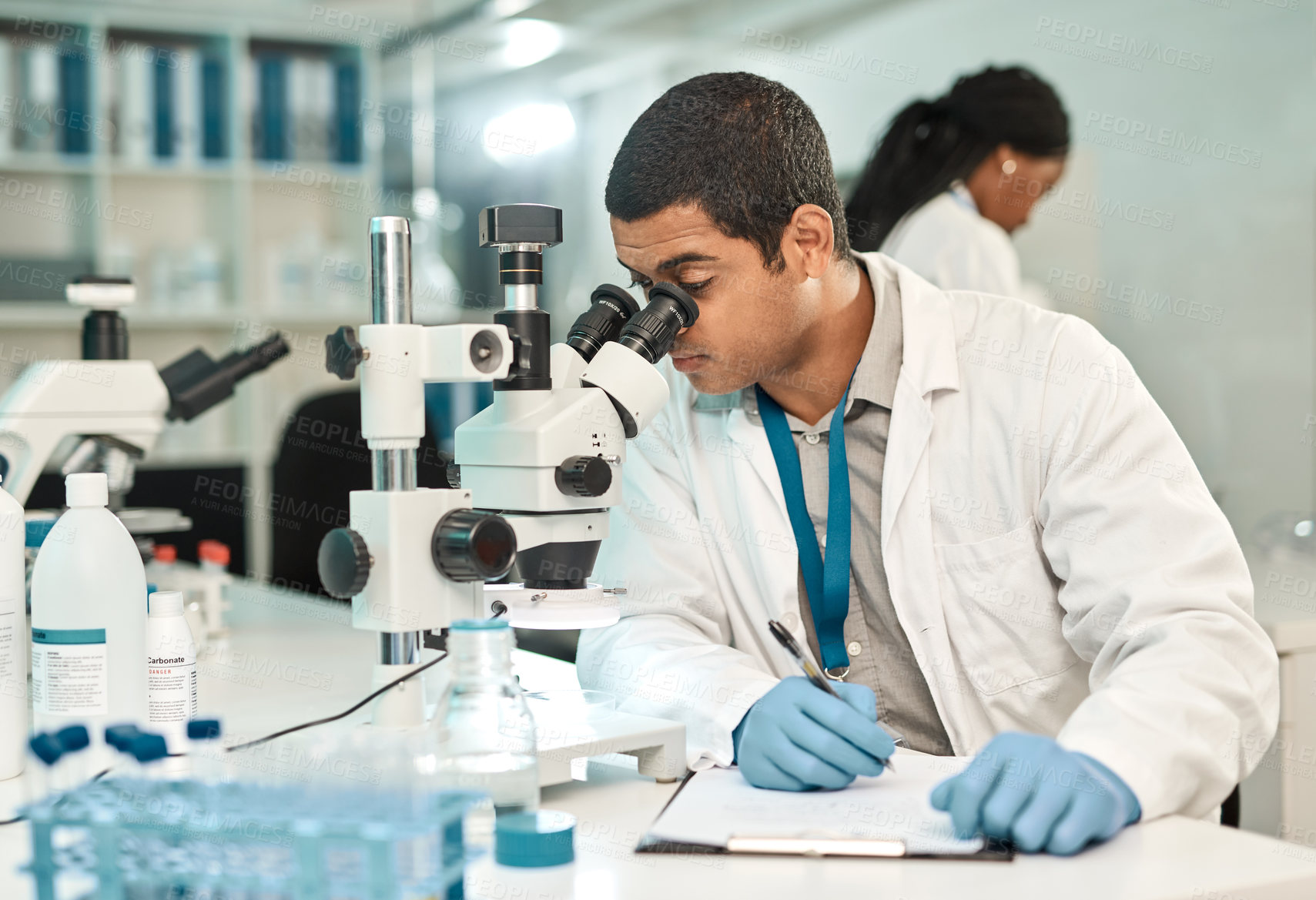 Buy stock photo Shot of a young scientist writing notes while using a microscope in a lab