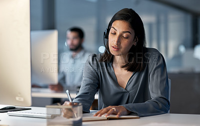 Buy stock photo Shot of a young woman using a headset and computer late at night in a modern office