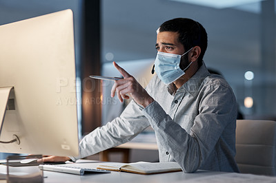 Buy stock photo Shot of a masked young man using a headset and computer late at night in a modern office