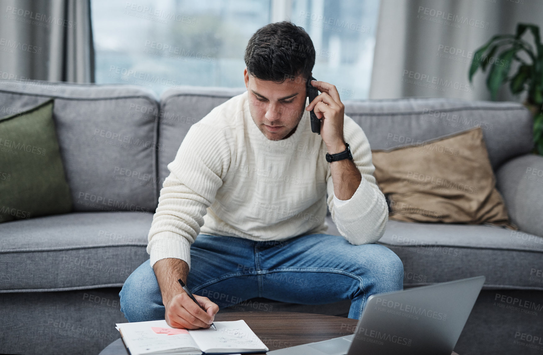 Buy stock photo Shot of a young man using a laptop and smartphone while writing in a notebook at home