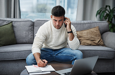 Buy stock photo Shot of a young man using a laptop and smartphone while writing in a notebook at home