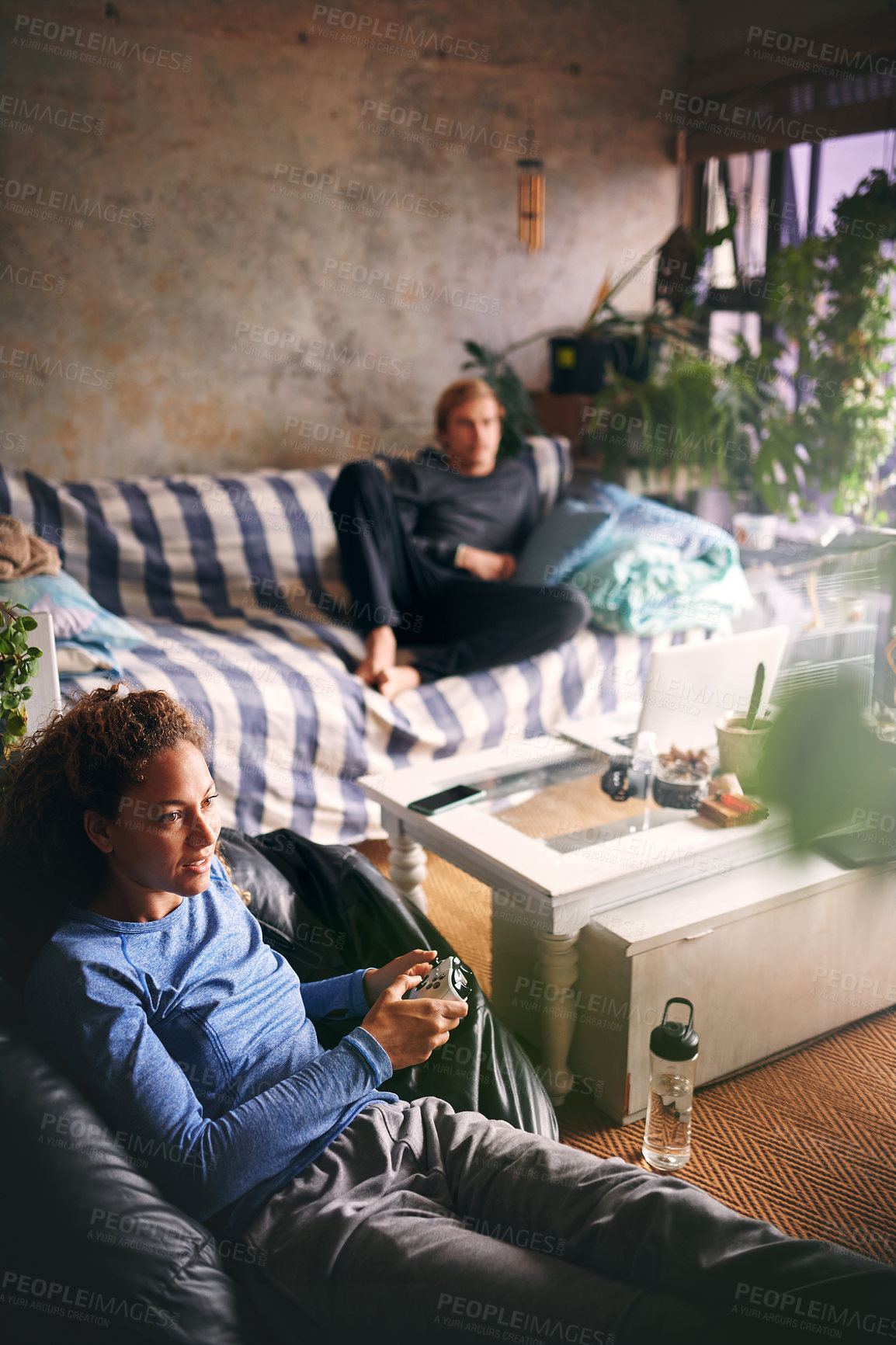 Buy stock photo Shot of a young woman playing video games while her boyfriend relaxes on the sofa in the background