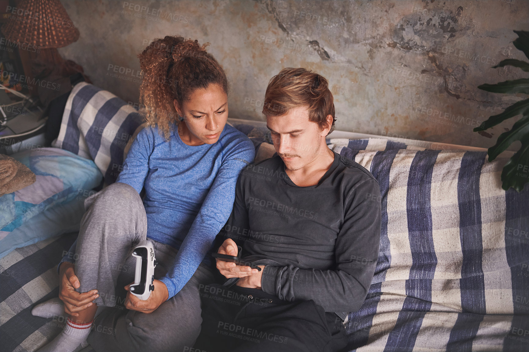 Buy stock photo Shot of a young couple using a smartphone while playing video games on the sofa at home