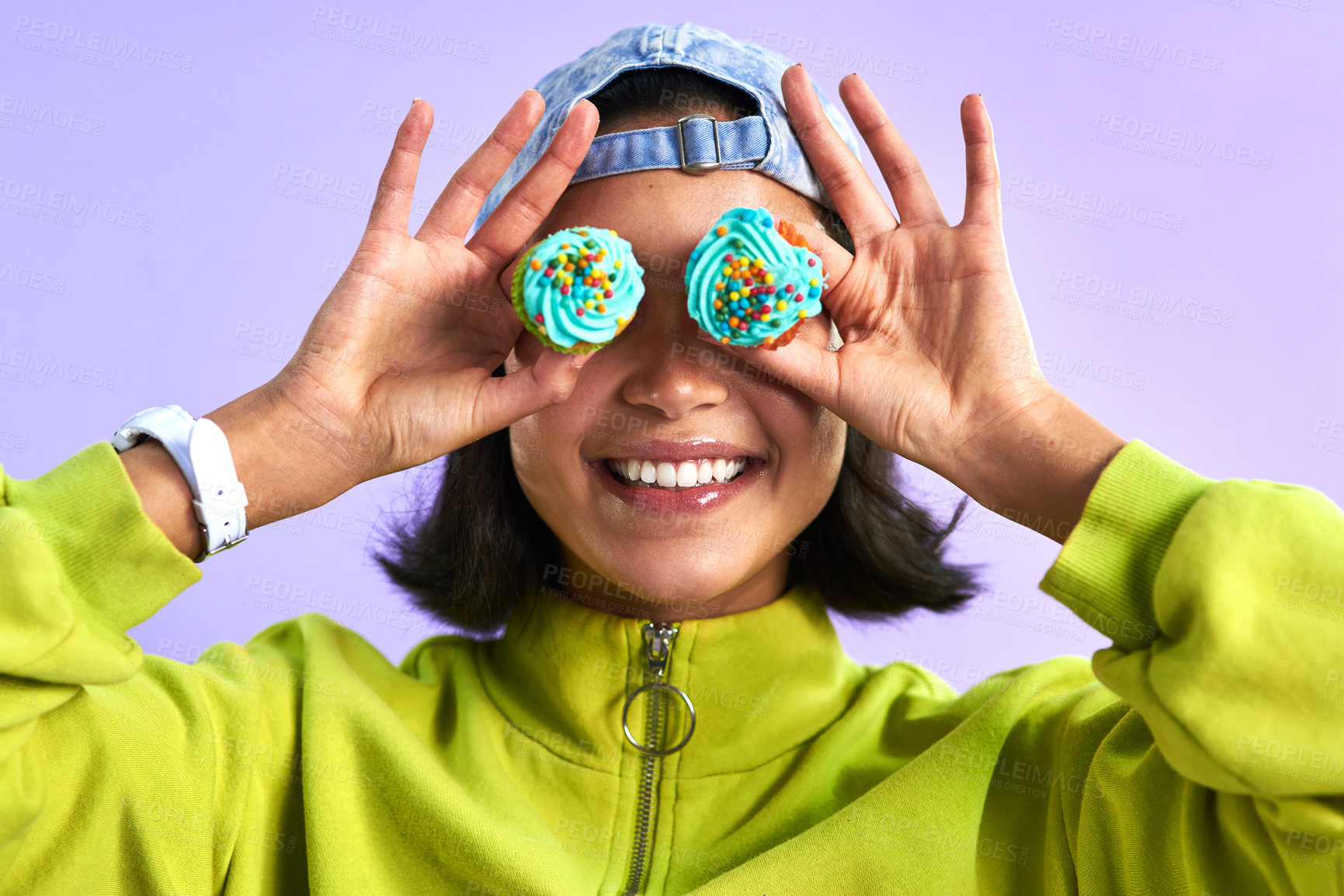 Buy stock photo Studio shot of a young woman holding cupcakes over her eyes against a purple background