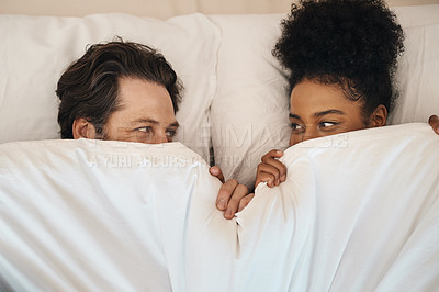 Buy stock photo Interracial, happy and relaxed couple lying in bed, bonding and looking shy while hiding after waking up together in the morning. Loving, carefree and romantic boyfriend and girlfriend being intimate