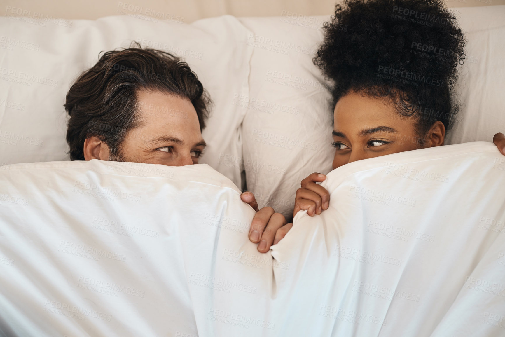 Buy stock photo Interracial, happy and relaxed couple lying in bed, bonding and looking shy while hiding after waking up together in the morning. Loving, carefree and romantic boyfriend and girlfriend being intimate