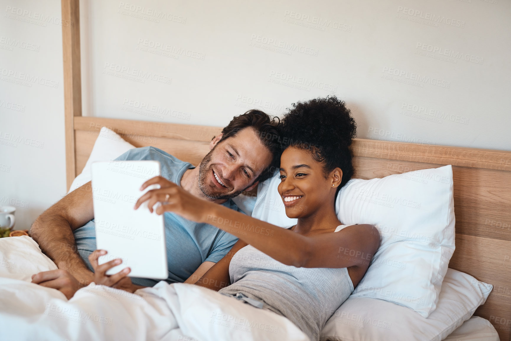 Buy stock photo Young, cozy interracial couple taking selfies on a tablet and lying together in modern bedroom at home. Smiling attractive woman relaxing with her handsome husband in the morning