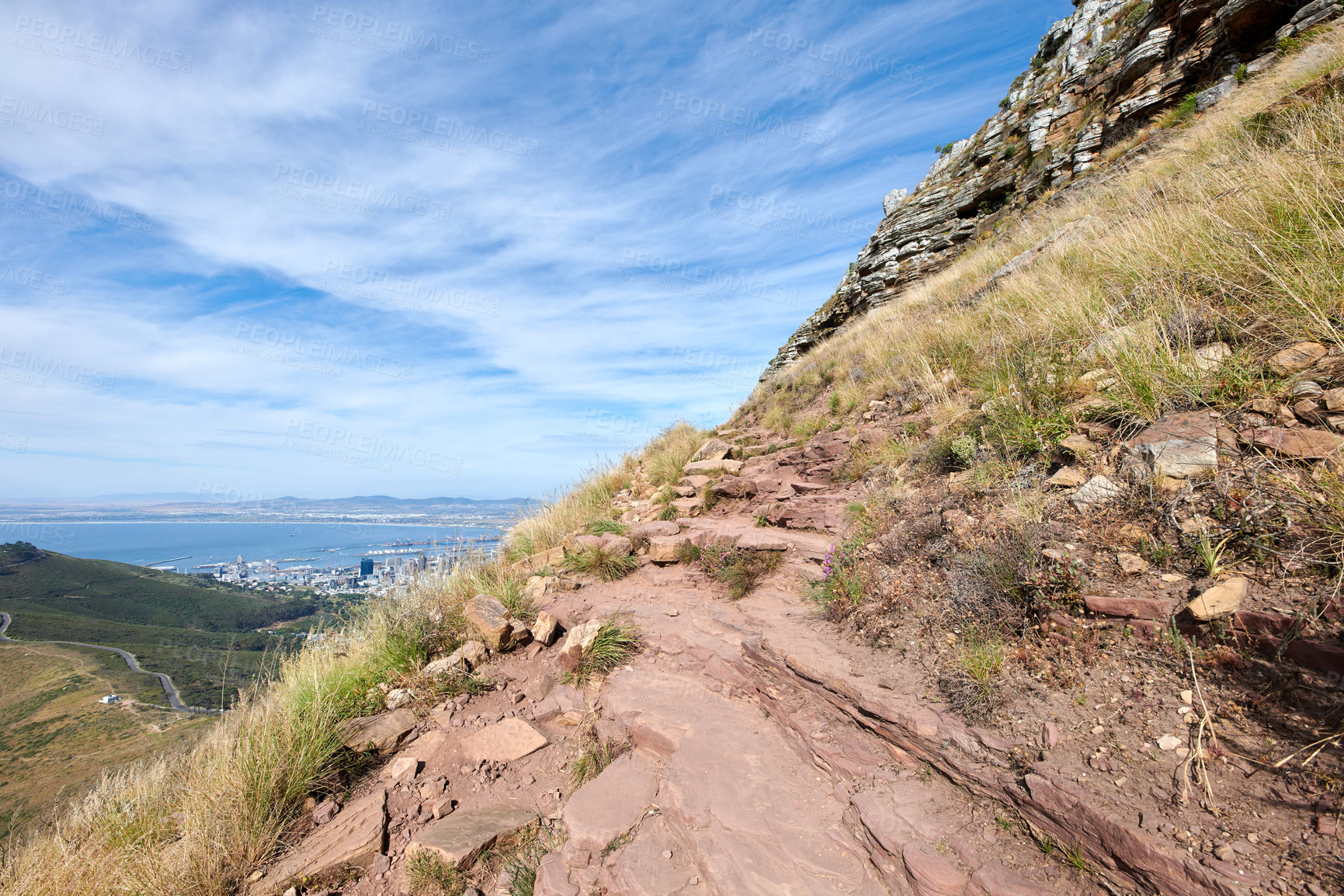 Buy stock photo Scenic of a rocky mountain slope with view of a coastal city by the sea and cloudy sky background with copyspace. Rugged landscape on a cliff with hiking trail on Lion's Head, Cape Town, South Africa