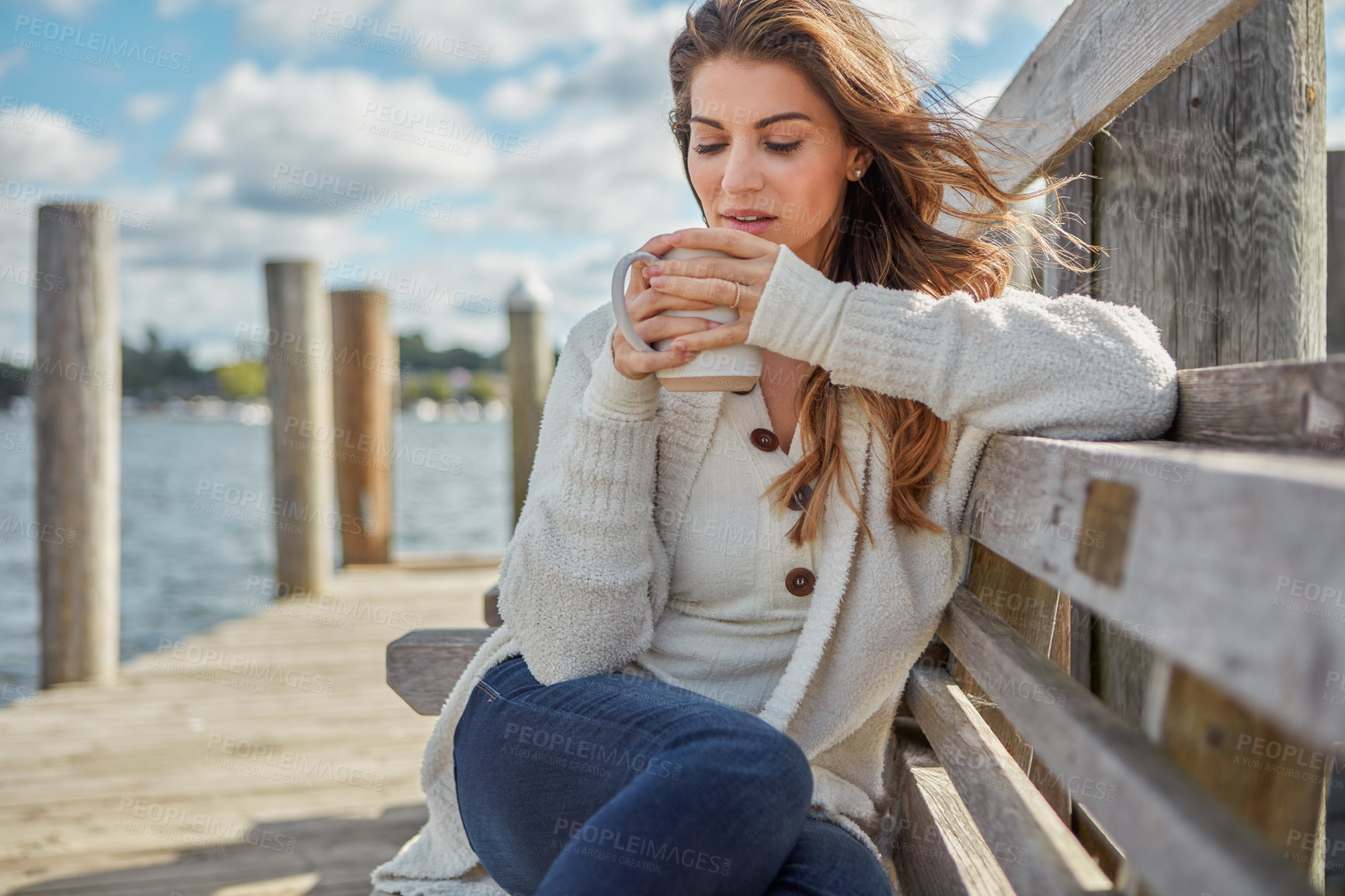 Buy stock photo Shot of a beautiful young woman enjoying a warm beverage while relaxing on a bench at a lake