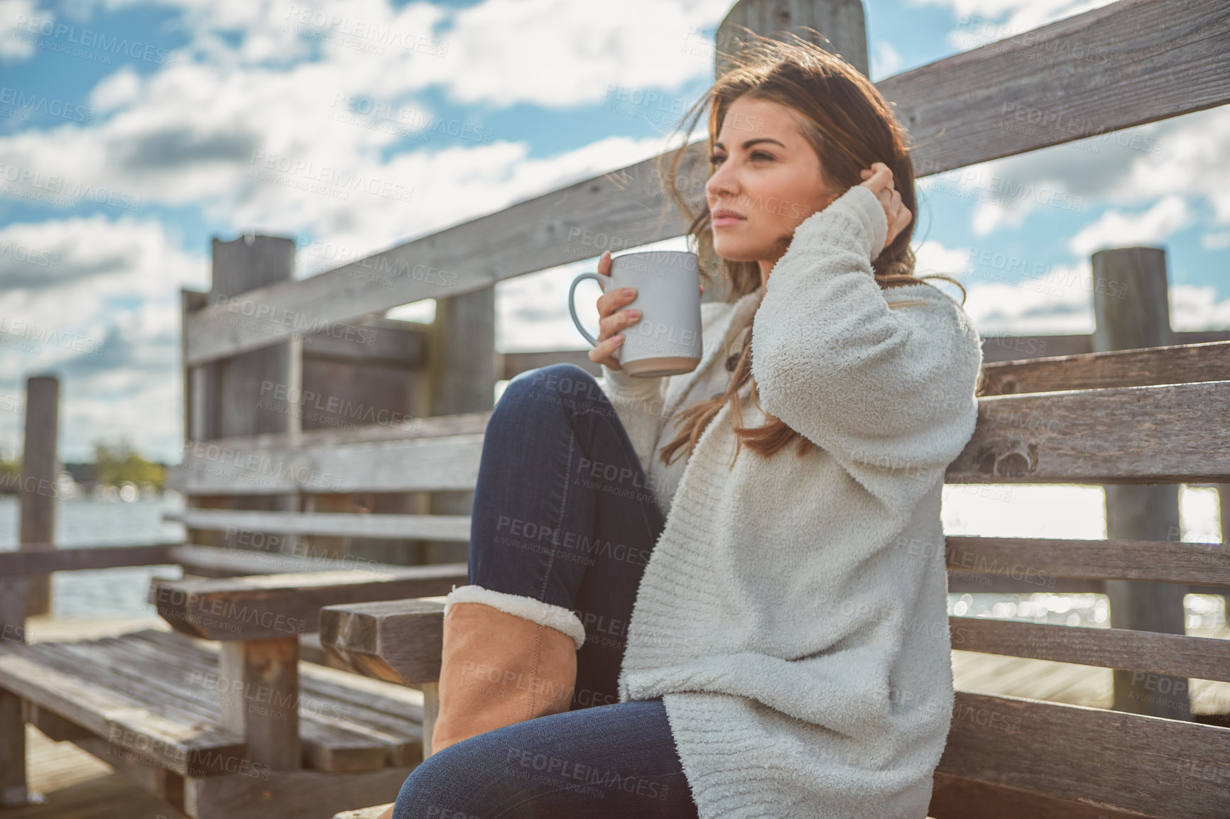 Buy stock photo Shot of a beautiful young woman enjoying a warm beverage while relaxing on a bench at a lake